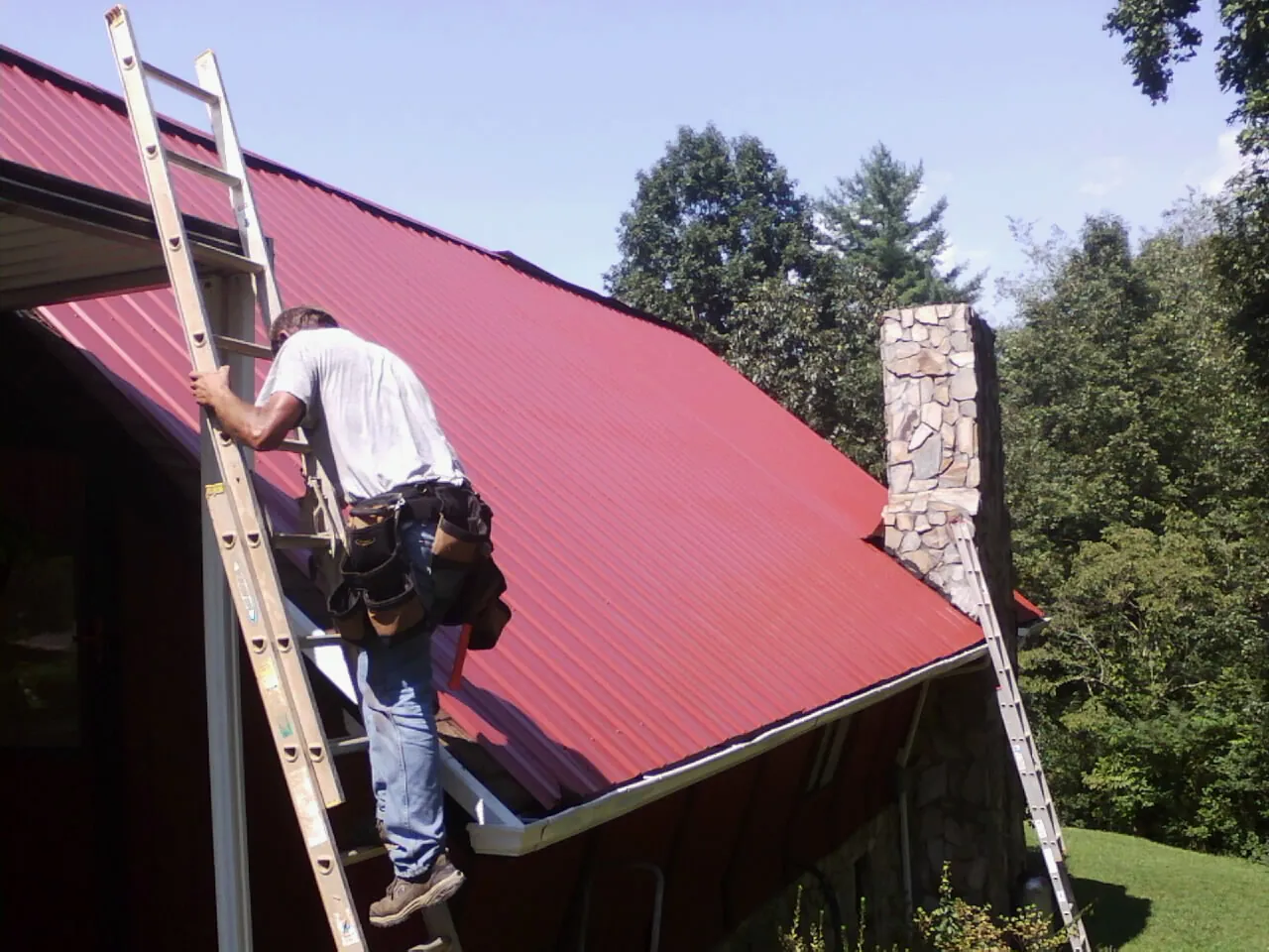 Roofer on a ladder beside a red metal roof and stone chimney — Soffit & Fascia Repair by Roofing Brothers in Goffstown